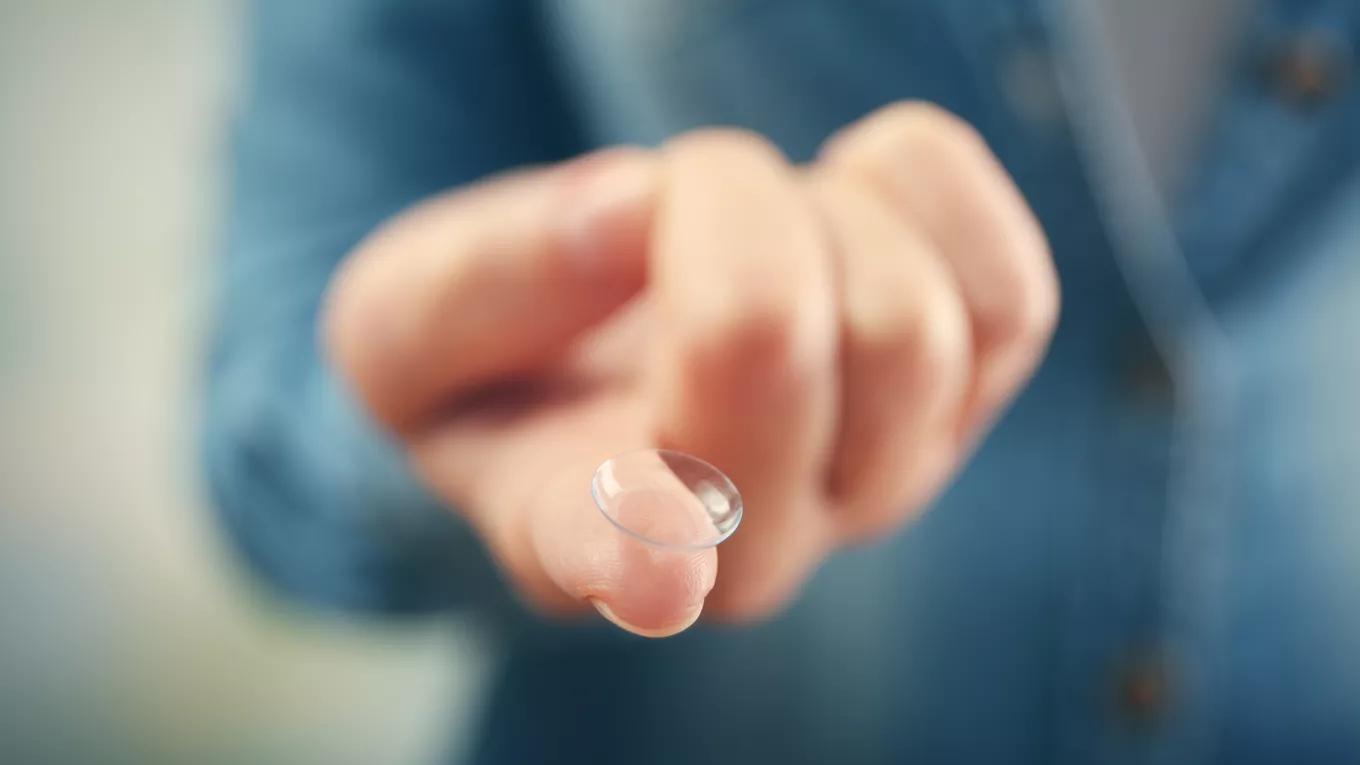 A close-up of a contact lens resting on an extended finger, with a blue sweater visible in the background