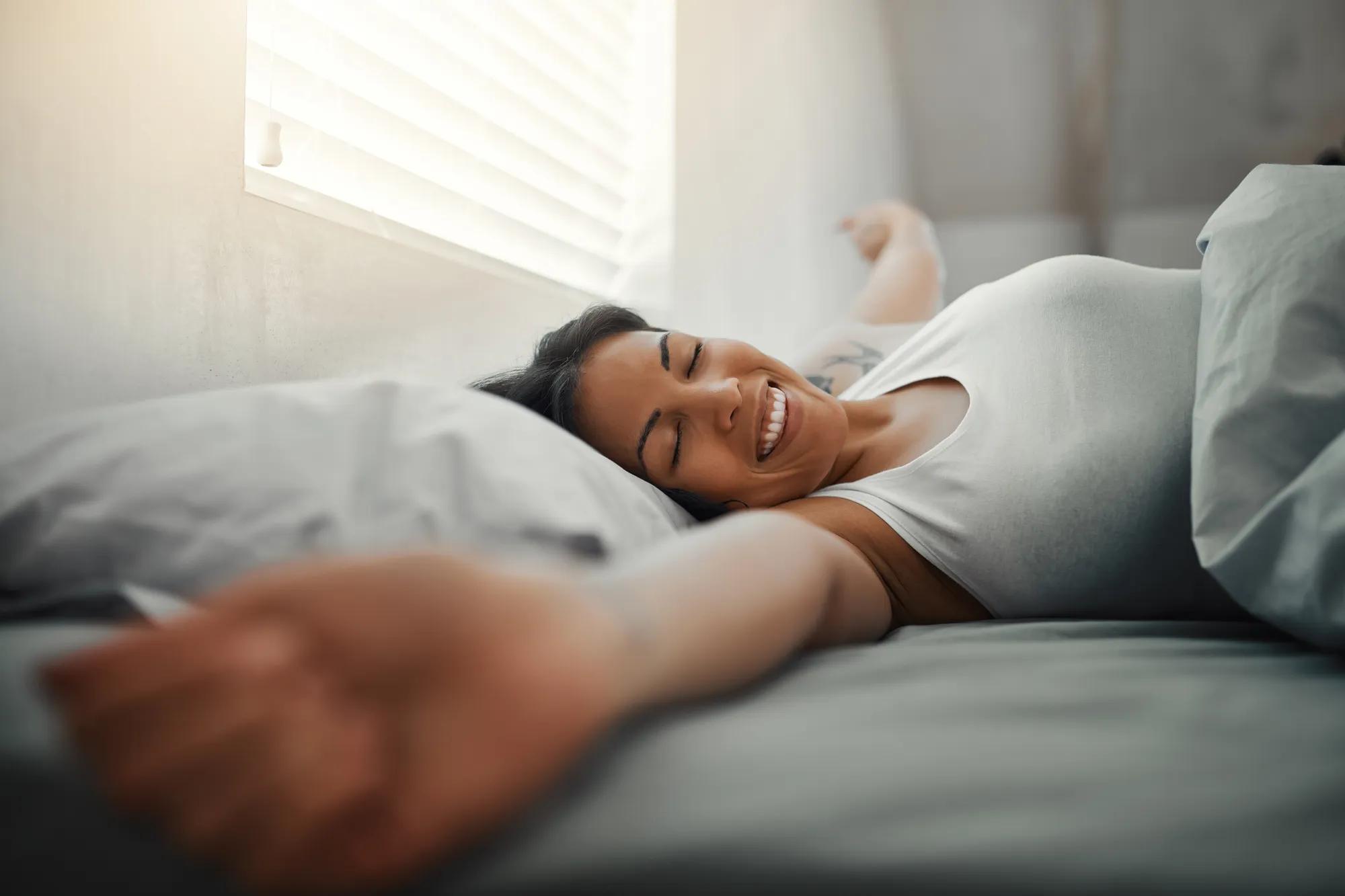 A woman in her bed stretches her arms wide and smiles as she wakes up and sun shines through her window