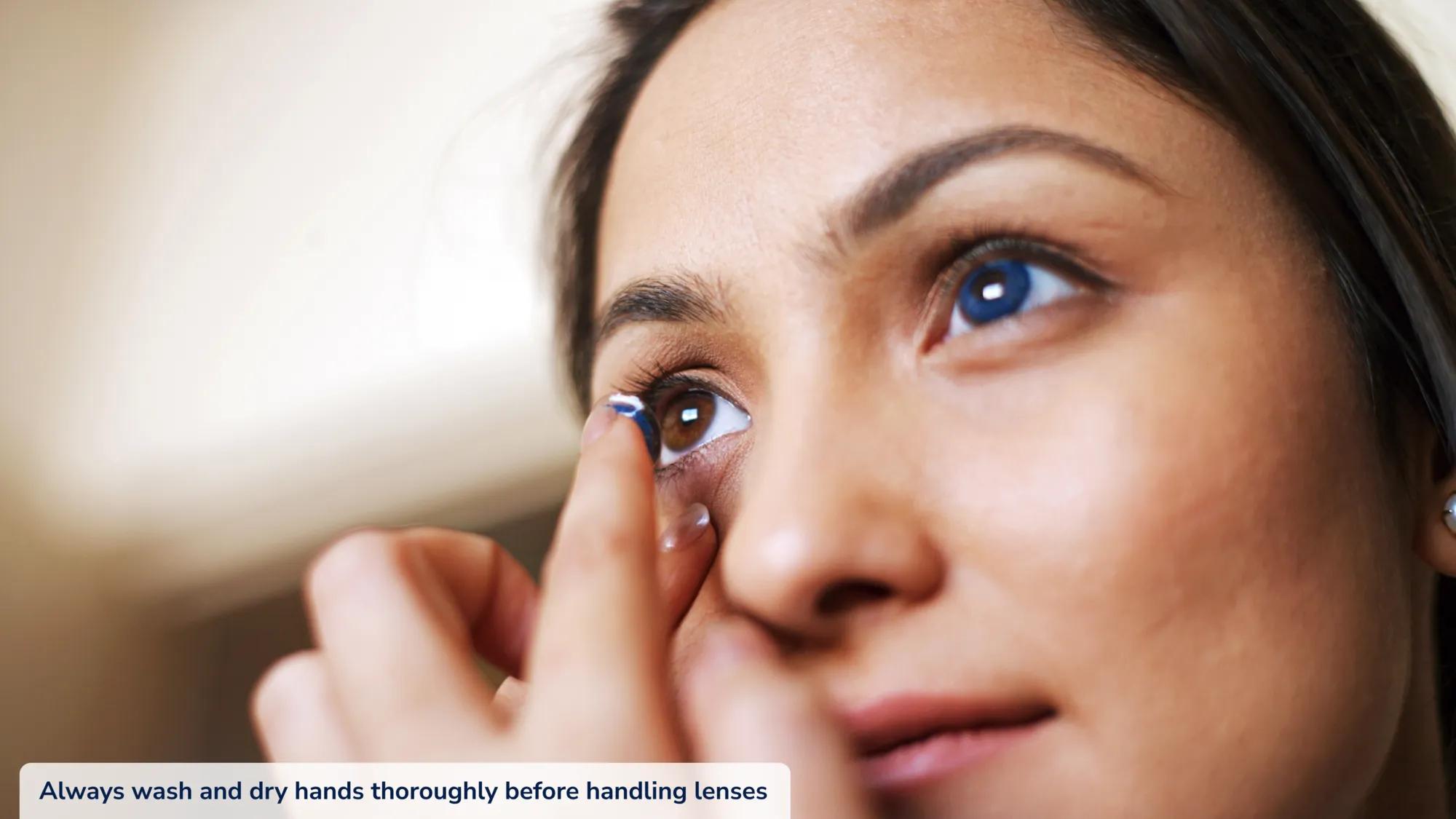 Close up of young woman with long dark hair placing a colored contact lens into her right eye