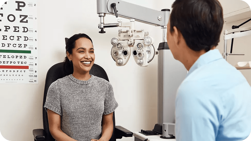 A woman in a grey shirt sits in a chair smiling and talking to her male eye doctor