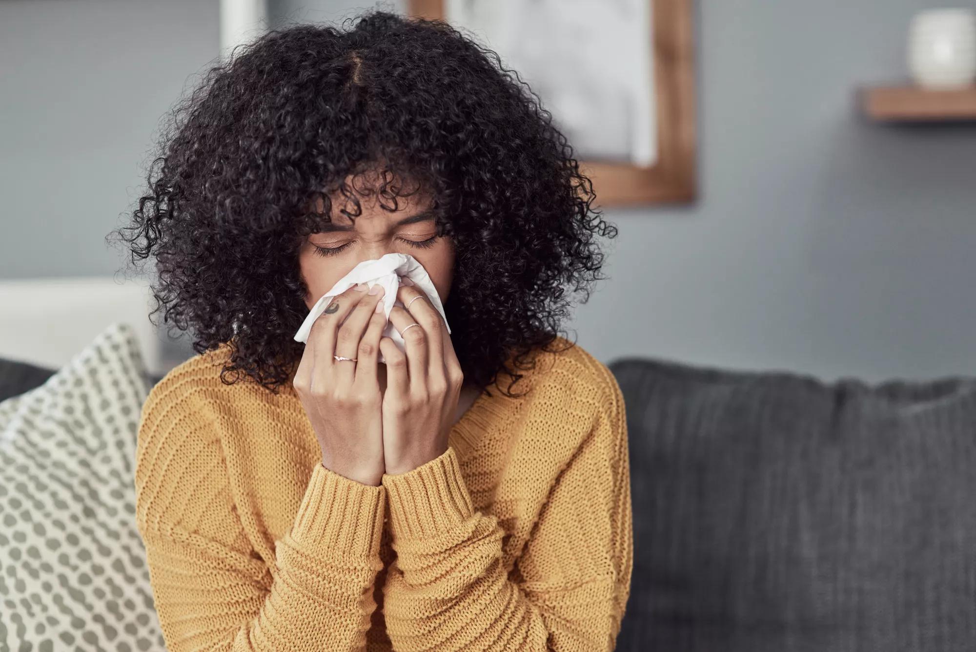 A woman with natural hair and a yellow sweater blows her nose into a tissue