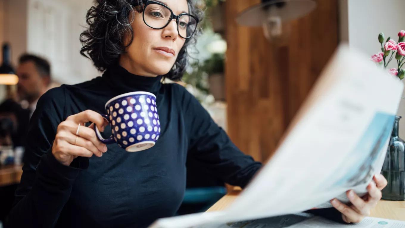 A woman wearing glasses sits reading a newspaper while she holds a blue coffee mug