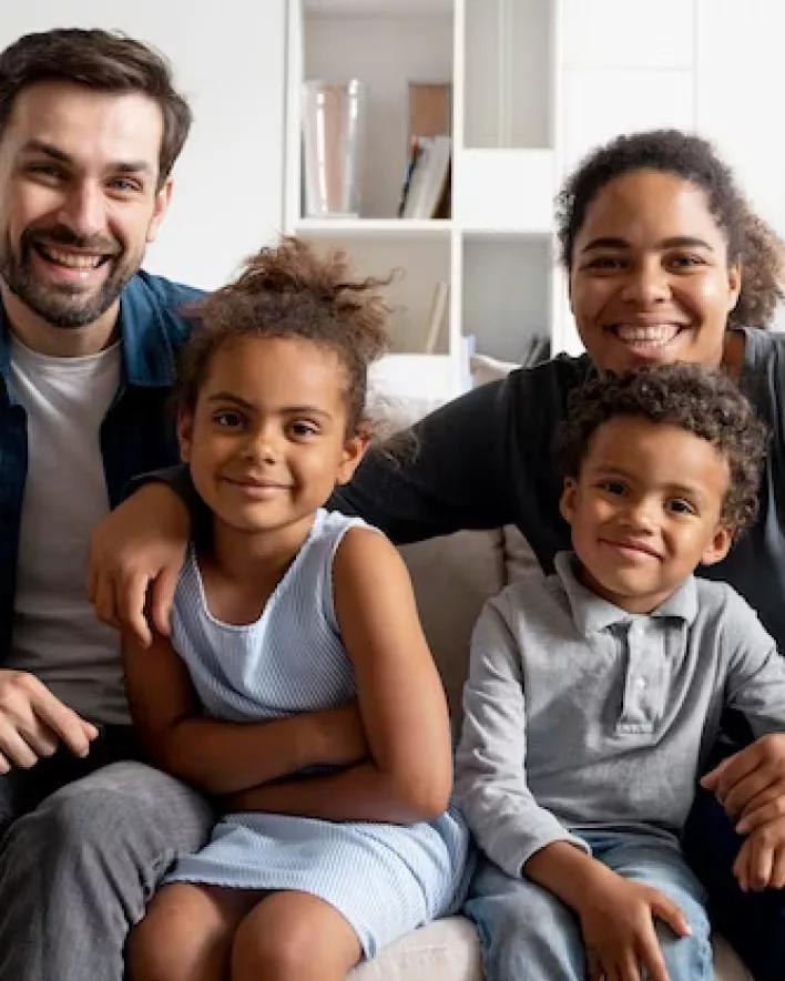 A smiling family of four sits together on a white couch with a bookshelf behind them.