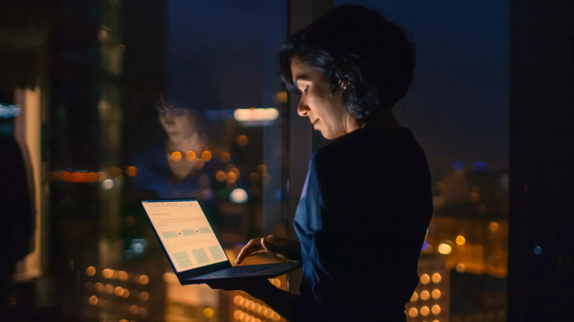 A businesswoman stands in front of a large glass window at night while working on a laptop