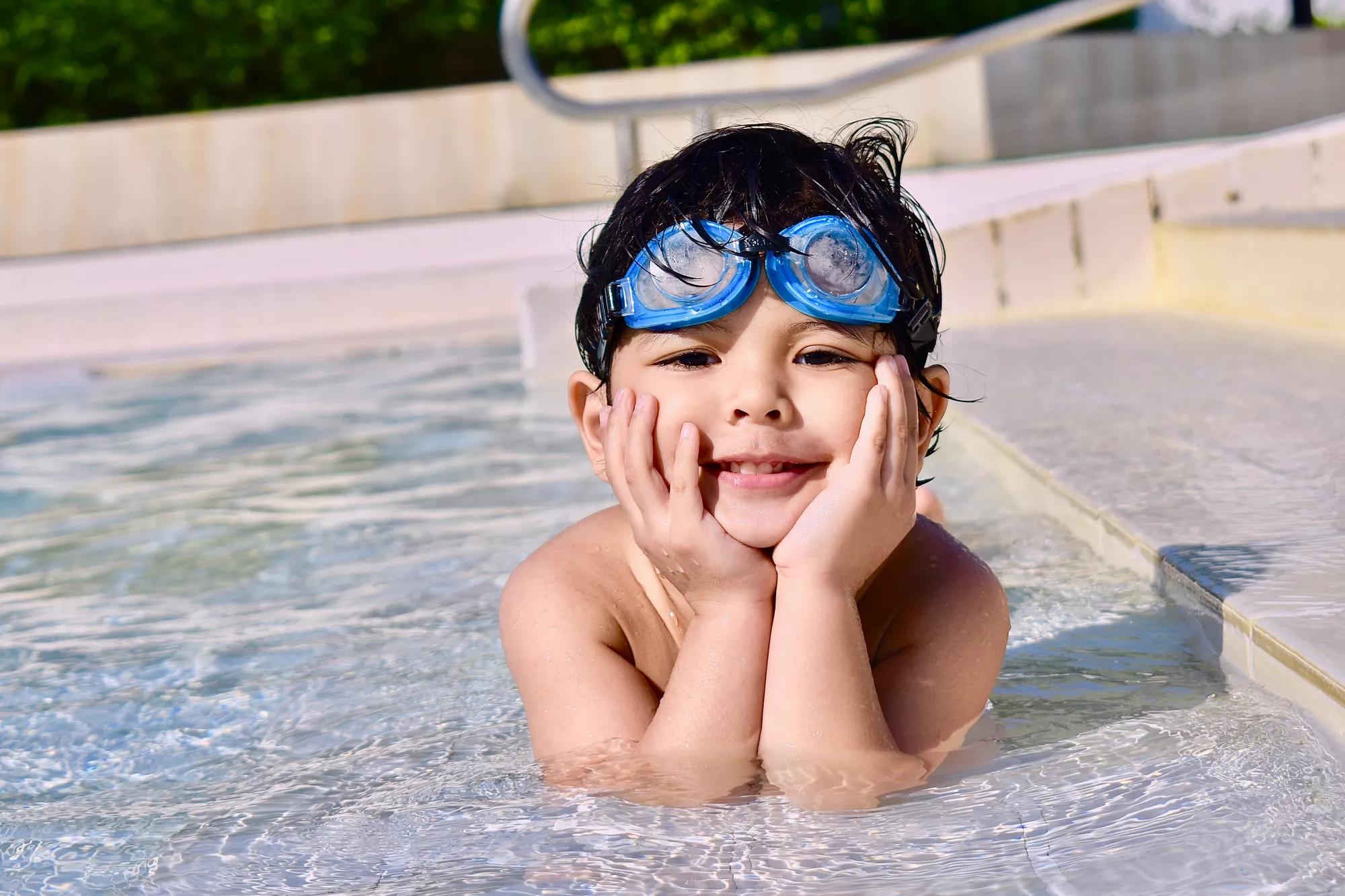 3-year old cute and handsome little boy lounges in the pool while smiling on a sunny day.