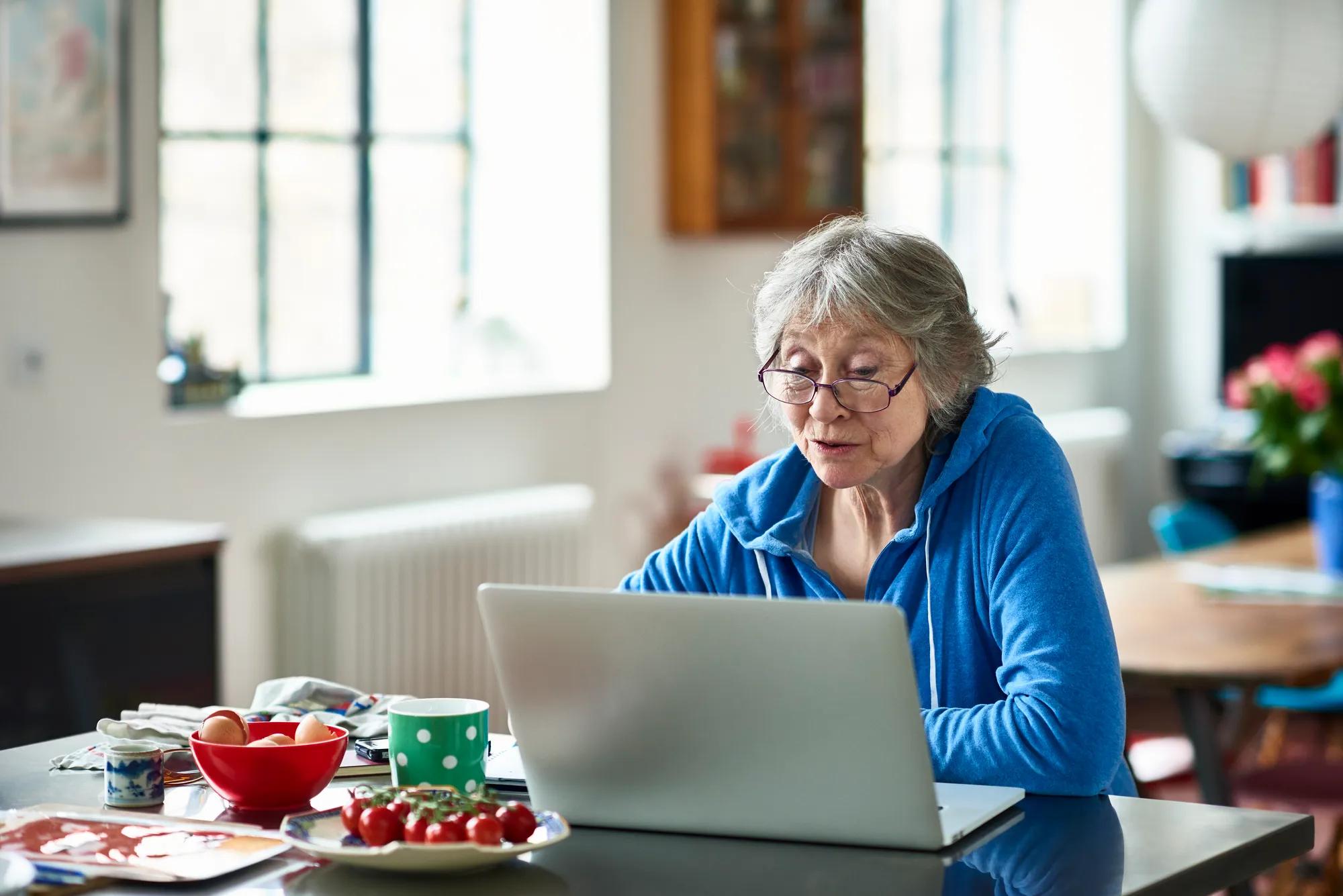 An older woman with grey hair and glasses sits at her kitchen island working on her laptop