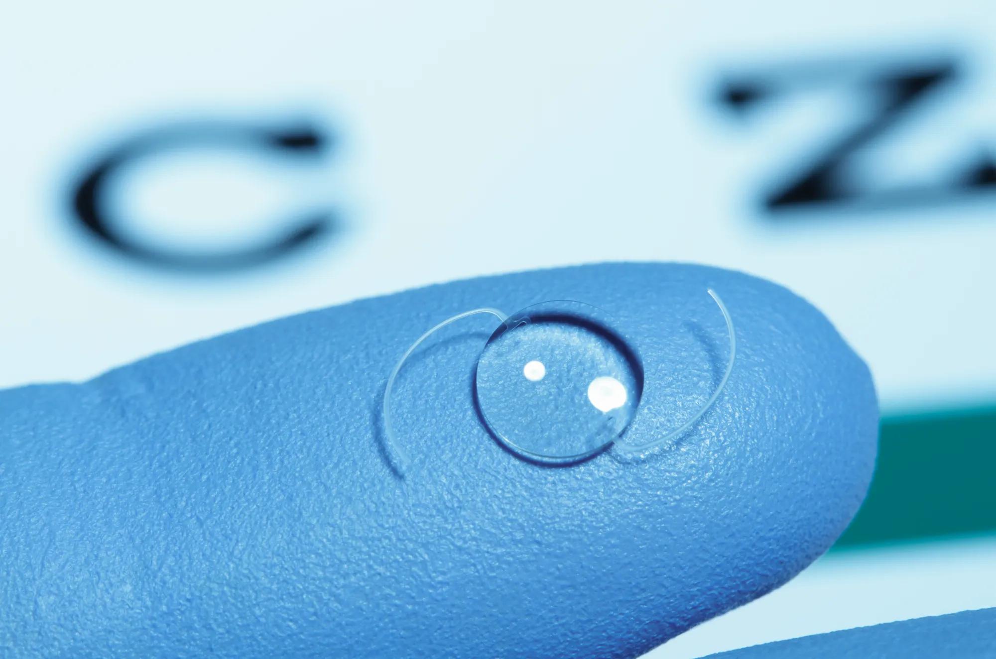 An intraocular lens on a gloved finger. Defocused Snellen eye chart is in the background. 