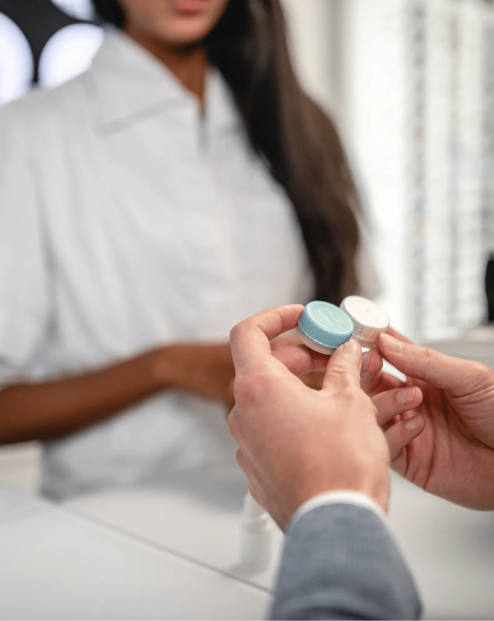 A woman with dark hair stands in the background as we see a close-up of a person holding a contact lens case