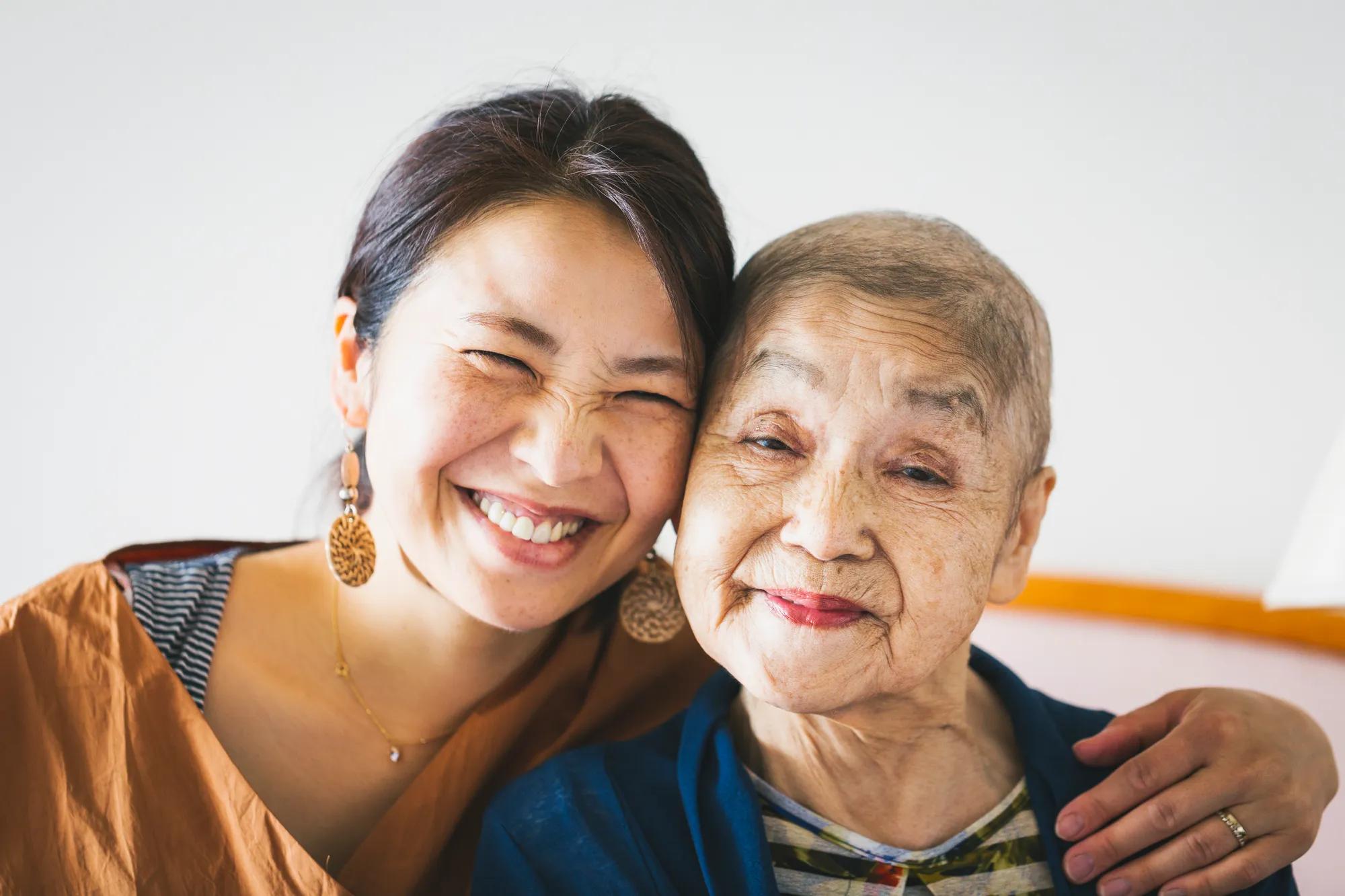 An Asian woman smiles and rests her head against her grandmother's head while putting her arm around her