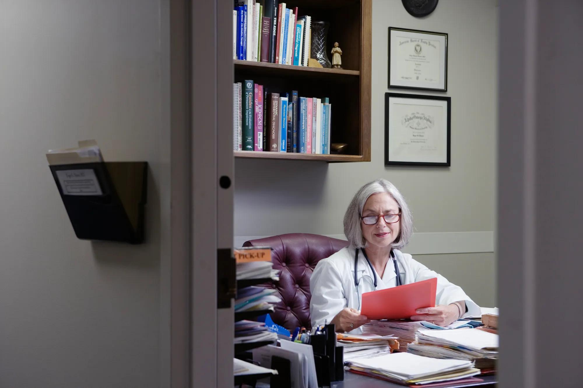 An older woman doctor wearing glasses sits at a desk reading a file