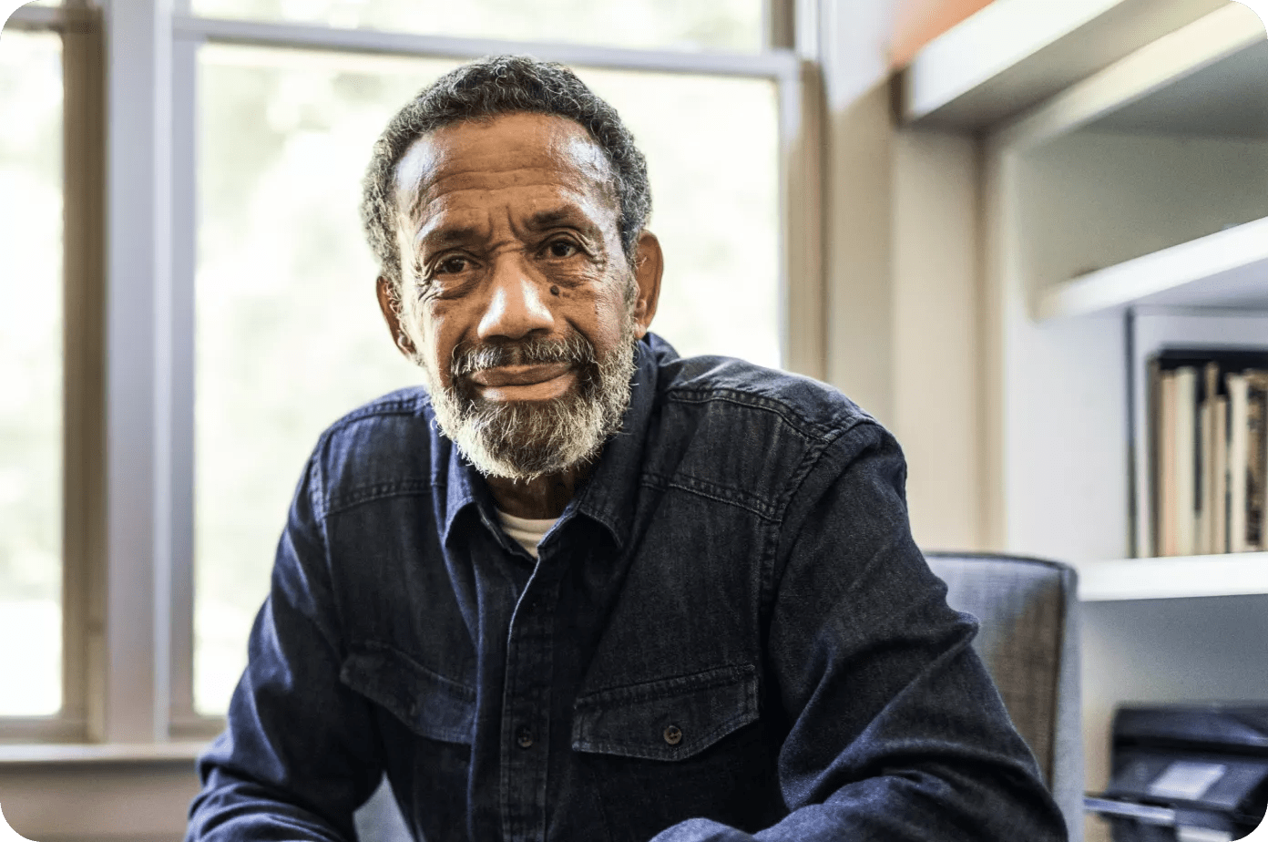 Older gray haired black man sitting by a window in front of a bookshelf