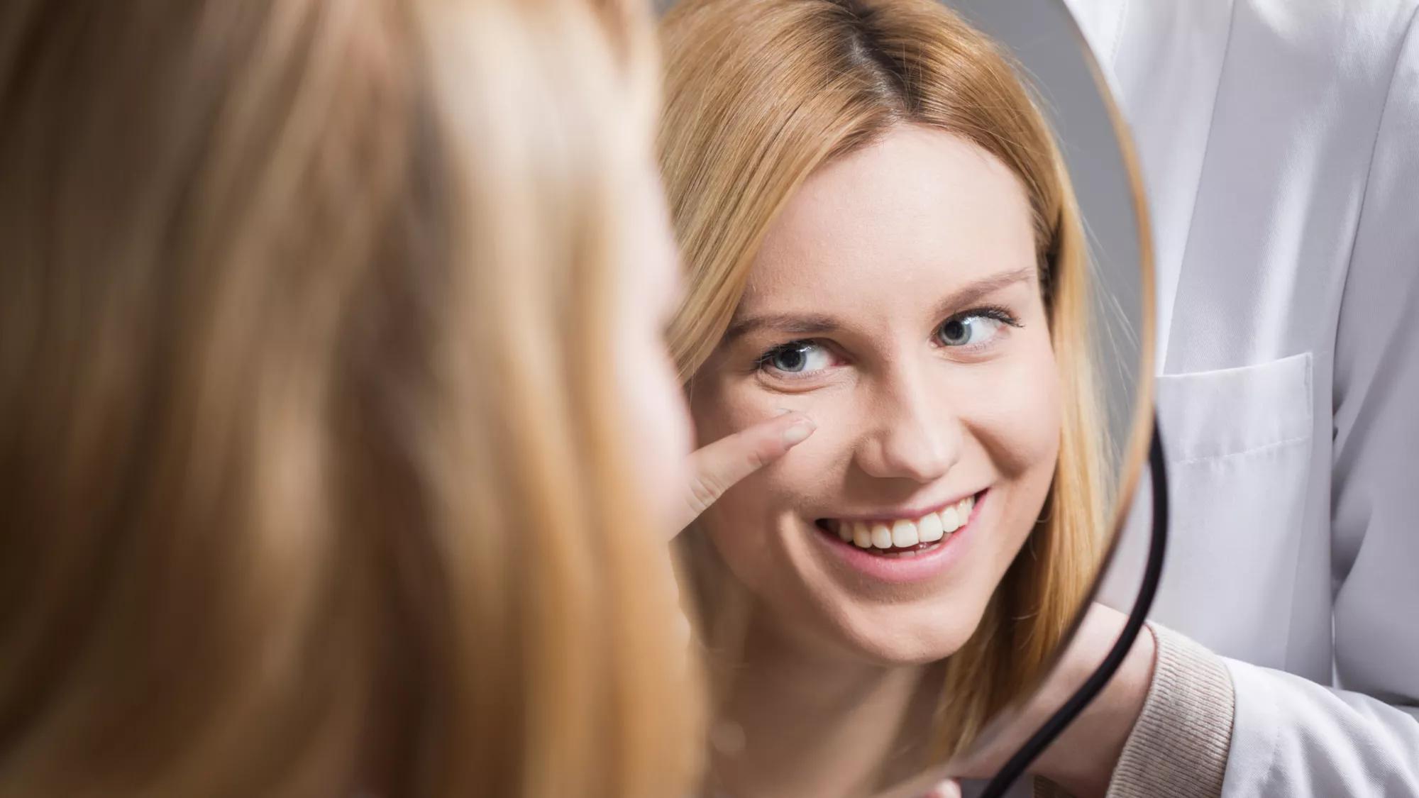 A smiling woman holding a contact lens on the tip of her finger while looking in a mirror