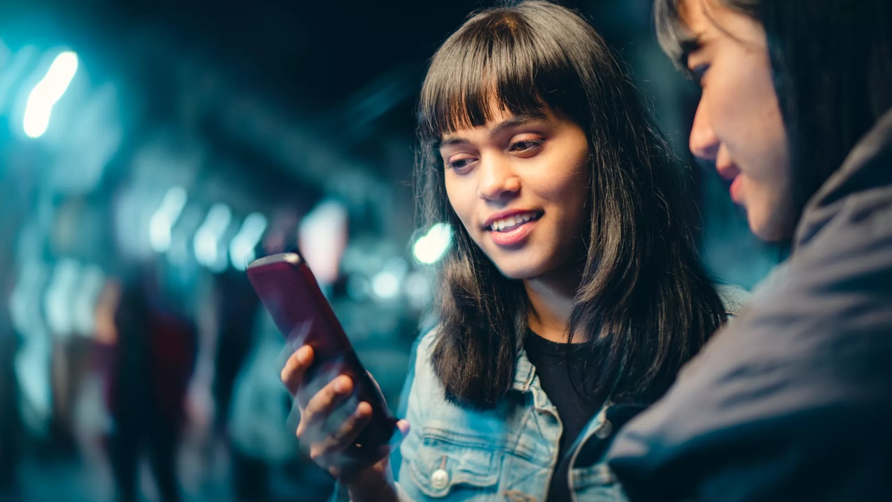 Outdoor image of happy Asian, Indian multiethnic female friends gossip and use smartphone together on city street at night