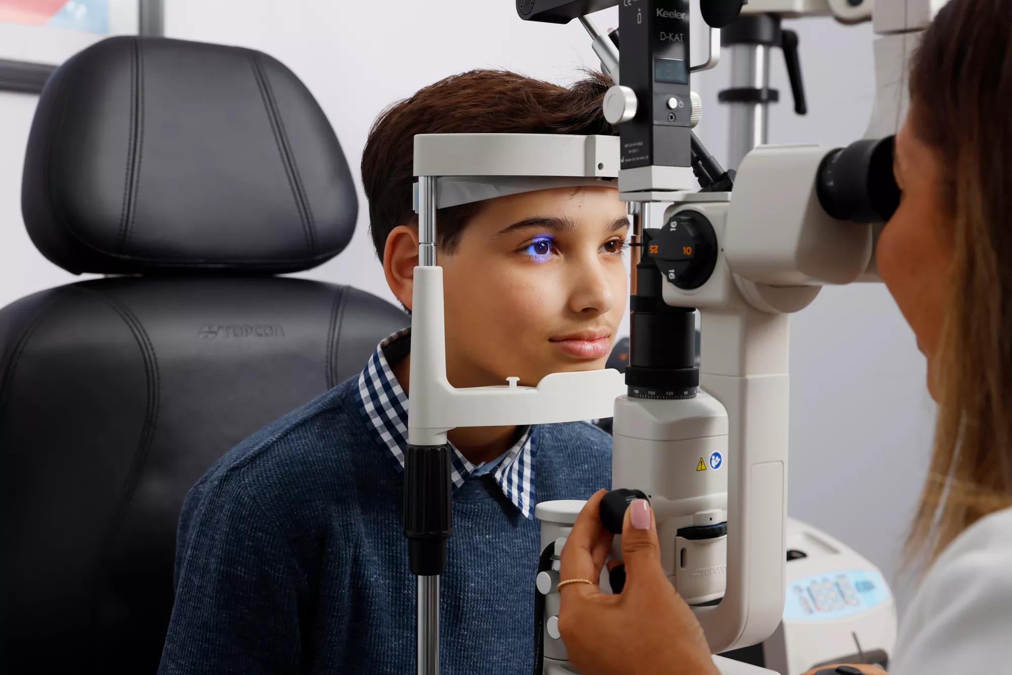 A young boy sits in a chair in front of an eye exam machine being operated by a female eye doctor