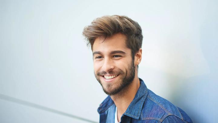 giovane ragazzo che sorride appoggiato ad un muro mentre indossa una camicia di jeans