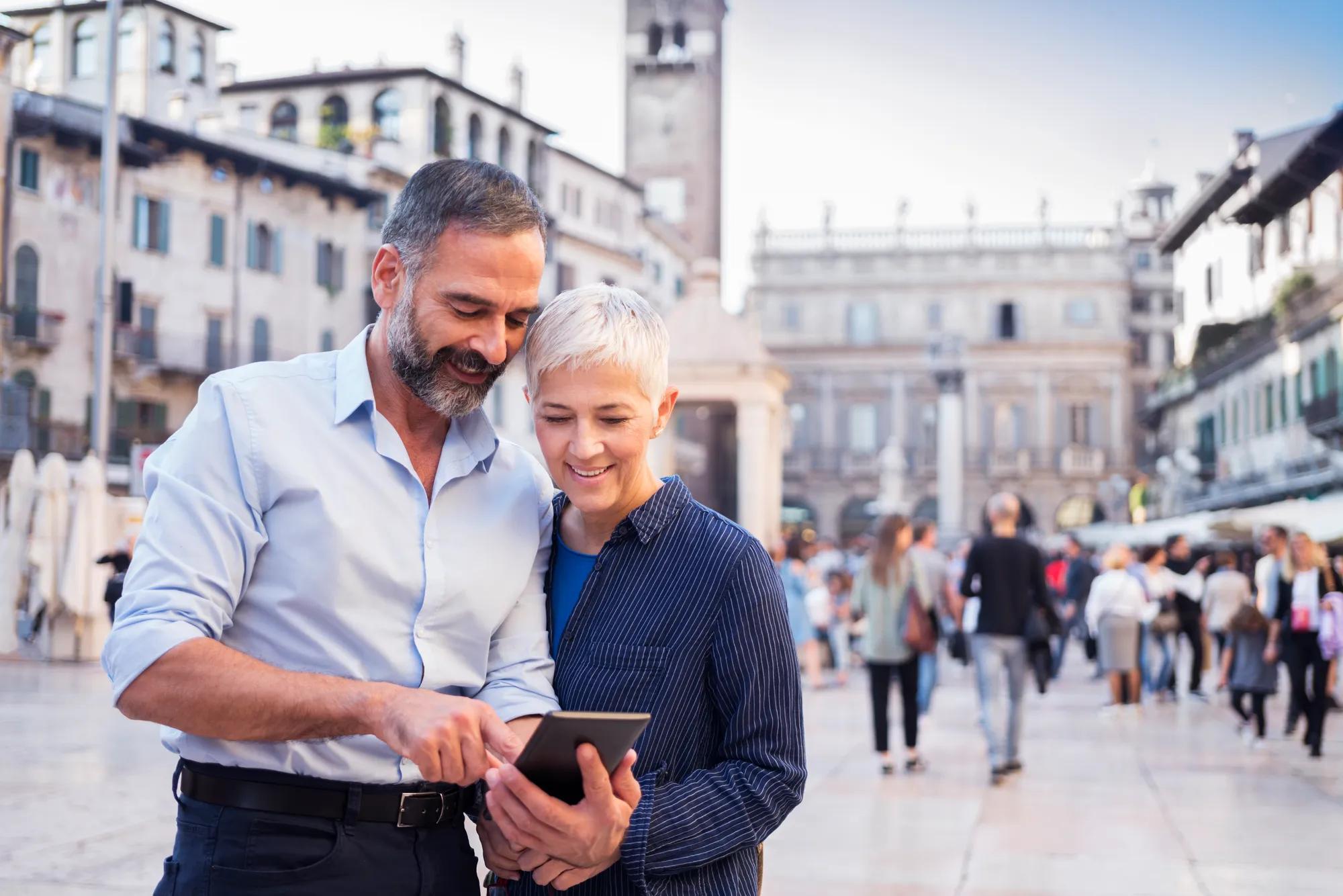 A man in a button-up shirt and his wife stand in a square looking at a phone in the man’s hand