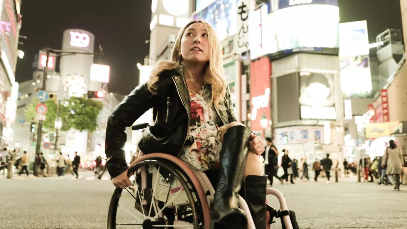 A woman sits in her wheelchair observing a brightly lit city at night