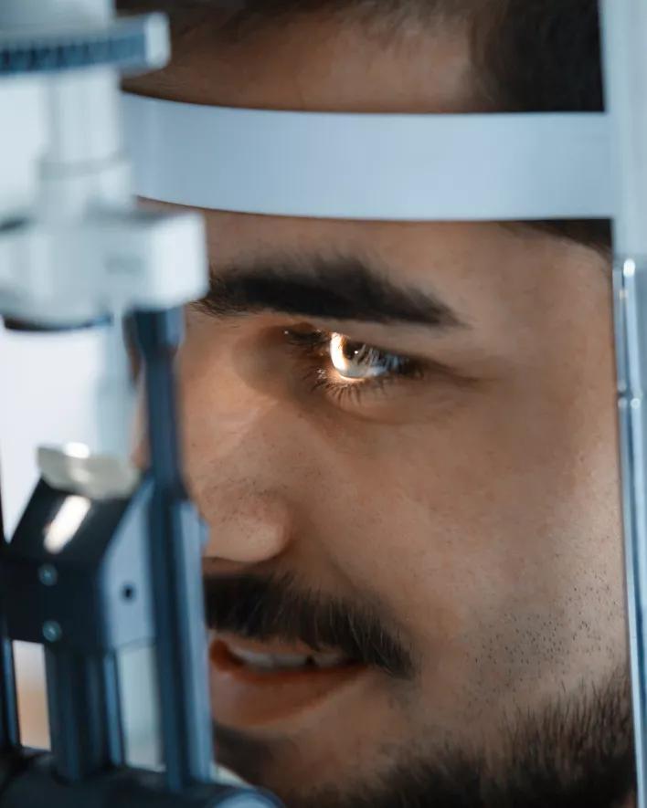 A man with a mustache and light shining in his eye has his face pressed up against an optometrist's machine