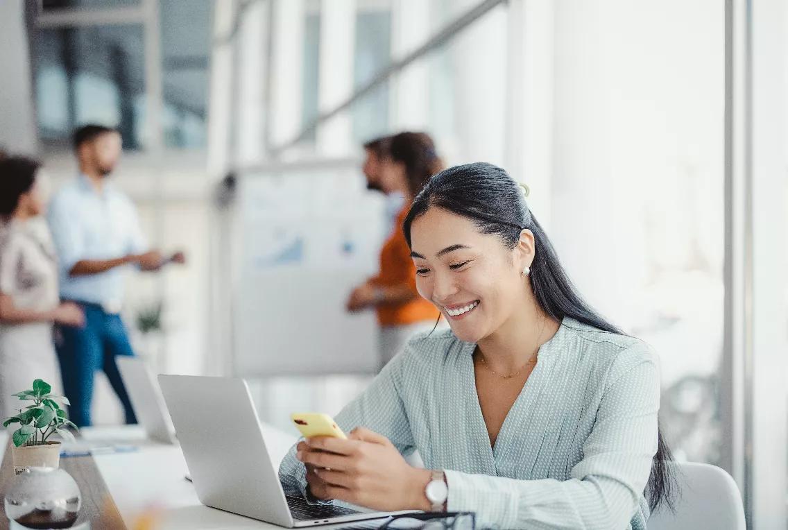 Young woman in front of a laptop looking at her cell phone with a smile.