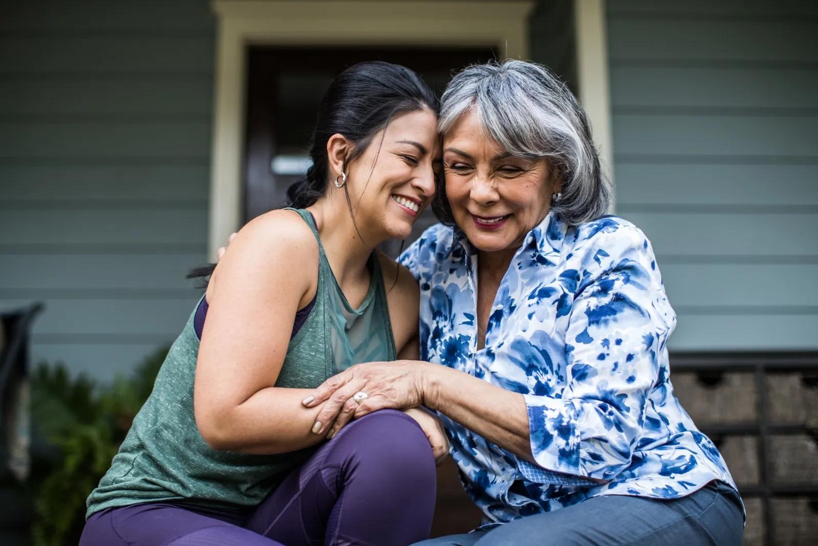 A woman with dark hair sits in front of her house embracing her mother in a floral shirt